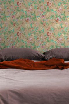 Cozy bedroom setup with a soft, floral wallpaper in peach and green tones, featuring a bed dressed in mauve and burnt orange bedding, alongside a hanging white pendant light. Wallpaper With Navy, Blue Grey Background, Frühling Wallpaper, Modern Bedroom Ideas, Wall Art Lighting, Spring Wallpaper, Floral Illustration, Orange Wallpaper