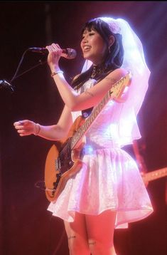 a woman in a white dress holding a guitar and singing into a microphone on stage