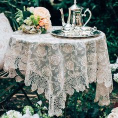 a tea set on a table in the middle of flowers and greenery, with an ornate lace tablecloth draped over it