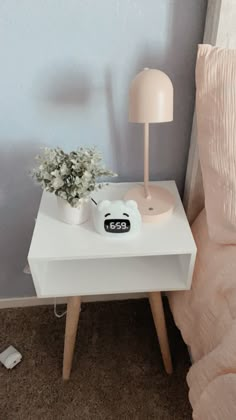 a small white table with a flower pot on it next to a lamp and a bed