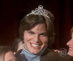 a smiling man wearing a tiara next to two other women in front of a red curtain