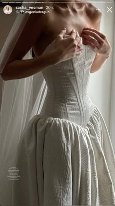 a woman in a white dress is standing by a window and adjusting her wedding dress