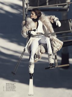 a woman sitting on top of a ski lift wearing white pants and a fur coat