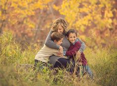 a woman and two children hugging each other in a field with fall foliage behind them