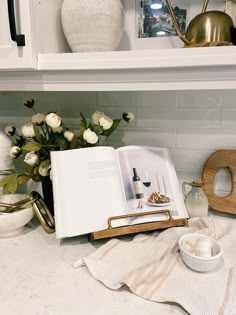 an open book sitting on top of a counter next to a bowl and vase filled with flowers