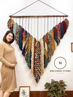 a pregnant woman standing in front of a wall hanging with multicolored yarns