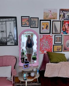 a woman standing in front of a mirror next to a pink chair and table with various pictures on the wall