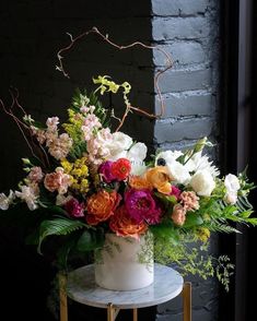 a white vase filled with lots of colorful flowers on top of a table next to a brick wall