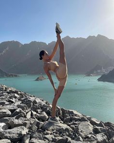 a woman is doing yoga on rocks near the water with mountains in the background and blue sky