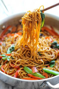 someone holding chopsticks with noodles and vegetables in a pan on the stove top