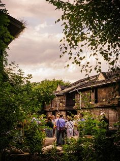 several people standing in front of an old wooden house surrounded by greenery and trees