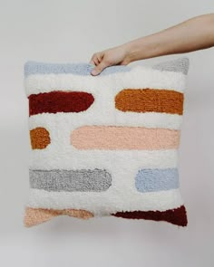 a hand holding a multicolored pillow on top of a white wall with a person's arm