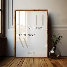 a white framed poster on a wooden floor next to a potted plant and window