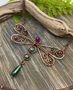 a dragonfly necklace with green and purple stones on it's back end, sitting on a piece of wood