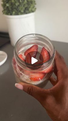 a hand holding a jar filled with strawberries on top of a table next to a potted plant