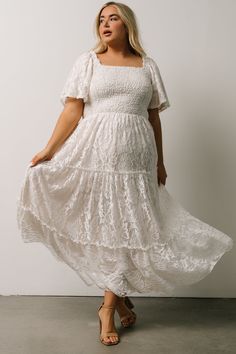 a woman wearing a white dress and sandals standing in front of a wall with her hands on her hips