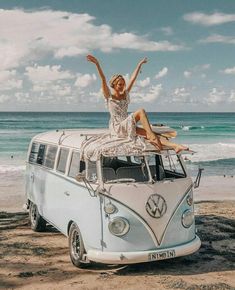 a woman sitting on top of a vw bus at the beach with her arms in the air