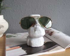 Moai glasses holder stand glasses stand concrete Moai statue unique gift concrete glasses holder desk decor Fathers days gift eyeglasses holder, sunglasses holder office decor. MATERIAL Concrete, sealant, pigment and paints. SIZE 70 x 90 x 122 mm HOW TO ORDER: 1. Select the colour. 2. Write down the gift message (optional). INCLUSIONS: Concrete glasses holder stand Eco-friendly packaging FREE gift message on the packaging (optional) PLEASE NOTE that due to the nature of handmade items, each order may slightly vary. There might be bubbles on the surface of the object but this is NOT a defect. We will endeavour to make sure these bubbles are kept to a minimum. The real color might look slightly different due to different screen setting. Each order will be made to order and posted out to you Eyeglasses Holder, Glasses Holder, Sunglass Holder, Eyeglass Holder, Aesthetic Room, Desk Decor, Eyewear Sunglasses, Fathers Day Gifts