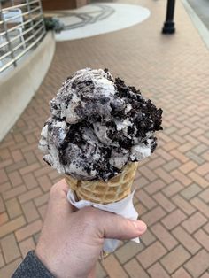 a person holding up an ice cream cone with chocolate chunks on it in front of a brick walkway