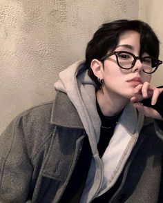 a person wearing glasses and a hoodie leaning against a wall with their hand on his chin