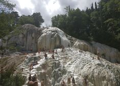 many people are swimming in the water near some rocks and trees, while others stand around them