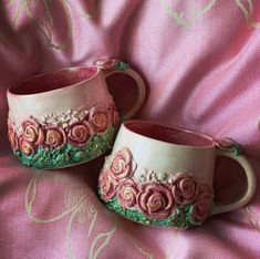 two coffee mugs sitting on top of a pink cloth covered tablecloth with roses painted on them