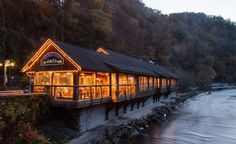 a restaurant on the side of a river at dusk with its lights on and windows lit up