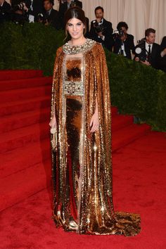 a woman in a gold gown and cape on the red carpet