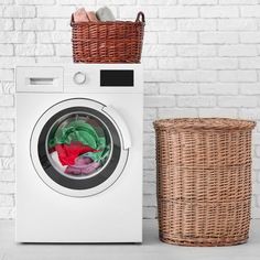 a washing machine with clothes in it next to a basket on the floor and a brick wall behind it