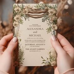 two hands holding up a wedding card in front of flowers and greenery on the background