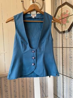 This pretty vest is made from permanent press fabric in a beautiful shade of medium blue. It has the original covered buttons and western style detailing on the back yoke. Very fitted. The measurements, taken with the vest lying flat, are: shoulder to shoulder, 12 1/2 inches; armpit to armpit, 15 inches; length, 22 inches in front and 22 inches in back; bottom edge, 17 inches. In very good condition. Blue Vest With Button Closure For Spring, Blue Cotton Vest With Buttons, Fitted Sleeveless Denim Vest With Button Closure, Classic Blue Cotton Vest, Vintage Fitted Denim Vest With Buttons, Fitted Blue Denim Vest With Buttons, Fitted Denim Vest With Button Closure For Work, Retro Fitted Denim Vest With Button Closure, Blue Denim Vest For Work With Buttons