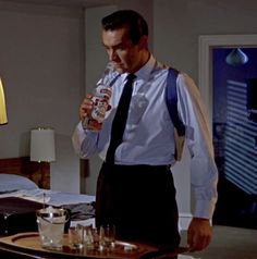 a man in suspenders drinking from a bottle while standing next to a table with glasses on it