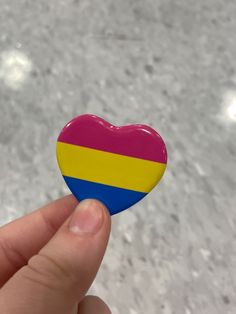 a hand holding a heart shaped pin with the colors of the flag of colombia on it