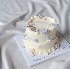 a three tiered cake with white frosting and purple flowers on the top is sitting on a card that says congratulations