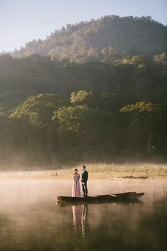 a man and woman standing on a boat in the middle of a lake with fog