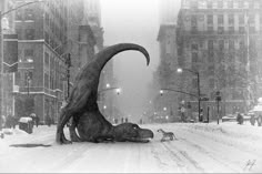 a large dinosaur laying on top of a snow covered street next to a cat walking in front of it