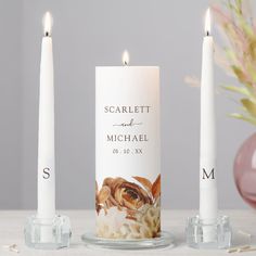 two white candles sitting next to each other on top of a table with flowers in the background