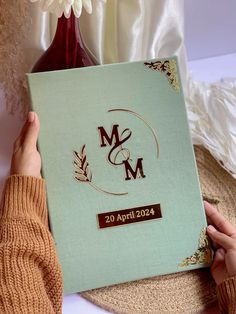 a person holding up a wedding album in front of a vase with flowers on it