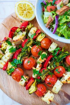 a wooden cutting board topped with lots of food next to a bowl filled with salad