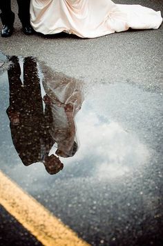 a bride and groom are reflected in a puddle on the ground as they walk down the street