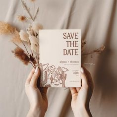 two hands holding up a save the date card next to some dried flowers and plants