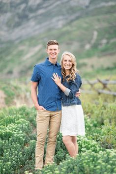 a man and woman standing next to each other in front of green bushes with mountains behind them