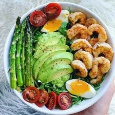 a person holding a white bowl filled with shrimp, avocado, tomatoes and asparagus