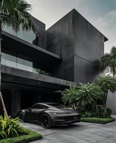 a black sports car is parked in front of a modern house with palm trees and bushes