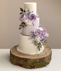 a three tiered wedding cake with purple flowers on the top and bottom, sitting on a tree stump