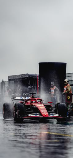 a red race car driving down a wet street next to a large black truck and some people