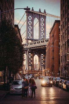 two people are standing on the sidewalk in front of cars and a bridge that is across from them