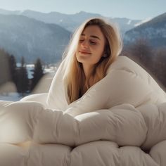 a woman laying on top of a bed covered in white sheets and blankets with mountains in the background