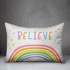 a white pillow with the words believe on it and rainbows in different colors against a gray background