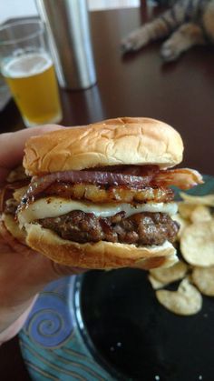 a hand holding a sandwich with meat and cheese on it next to chips in front of a glass of beer